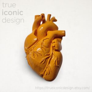 Scultura di cuore anatomico giallo ocra con dettagli intricati delle vene. La scultura ha una finitura metallica ed è posta su uno sfondo bianco. Il testo "true iconic design" è visibile in alto a sinistra.