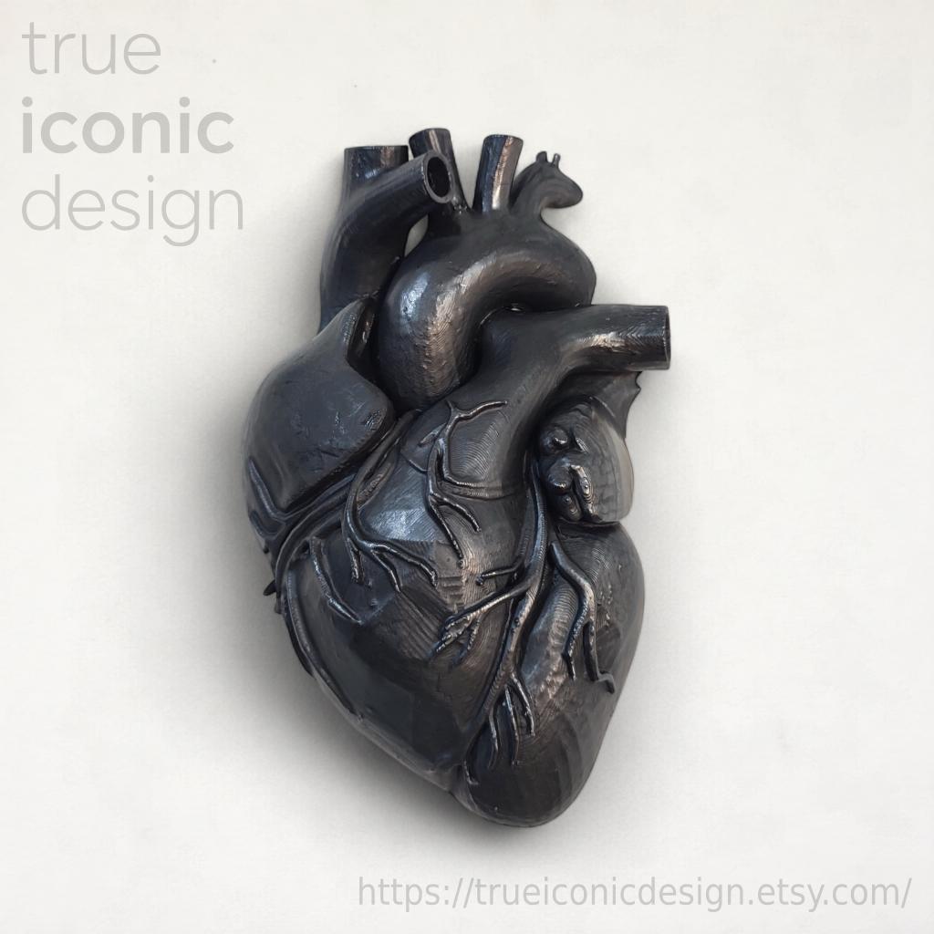Scultura di cuore anatomico grigio scuro con dettagli intricati delle vene. La scultura ha una finitura metallica ed è posta su uno sfondo bianco. Il testo "true iconic design" è visibile in alto a sinistra.